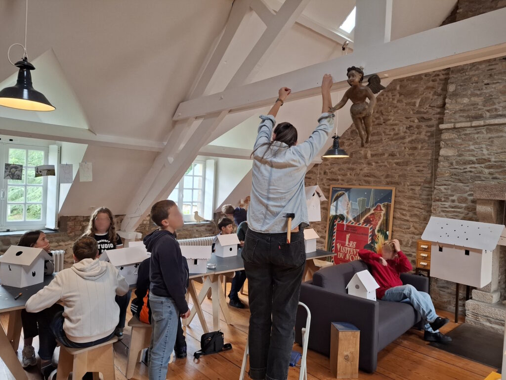 L'artiste Julie Chapallaz aide un élève à suspendre sa maquette de maison à une poutre de l'atelier de la Maison Jacques Prévert
