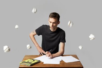 Un homme est assis à une table, il cherche l'inspiration pour écrire. Autour de lui pleuvent des boulettes de papier.