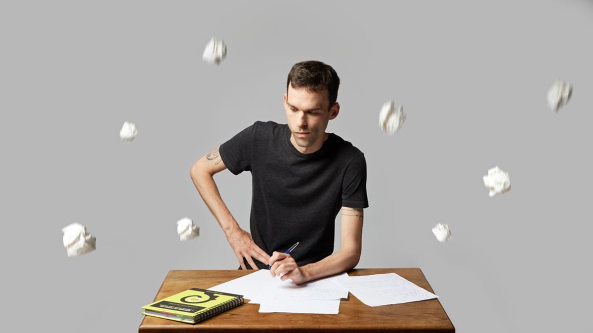Un homme est assis à une table, il cherche l'inspiration pour écrire. Autour de lui pleuvent des boulettes de papier.