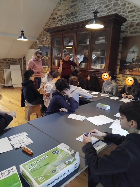Dans une pièce intérieure, des enfants sont assis à une table avec des feuilles devant eux. Ils sont tournés vers une femme qui est debout et leur parle. A sa gauche se trouve une autre femme. Derrière elles, il y a une grande bibliothèque.
