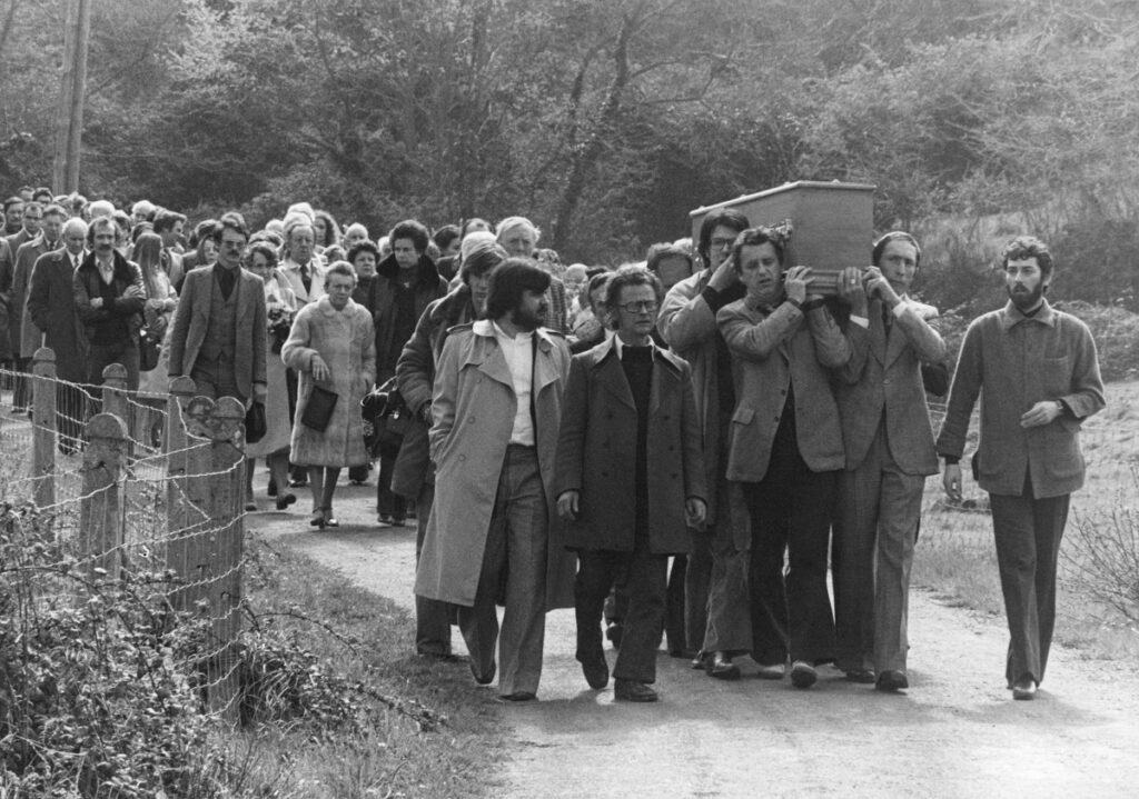Trois hommes portent un cercueil sur une route de campagne. Ils sont suivis par un cortége funéraire.