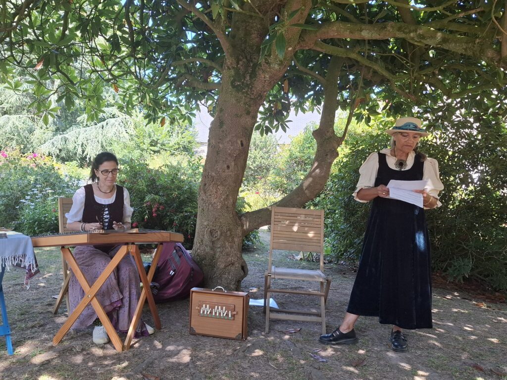 Une musicienne joue de l'instrument dulcimer. A ces côtés, debout, une femme lit un texte à voix haute.