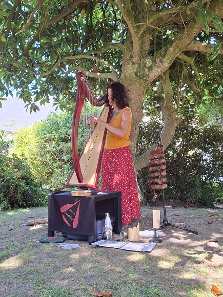 Une harpiste joue devant un arbre, dans un jardin.